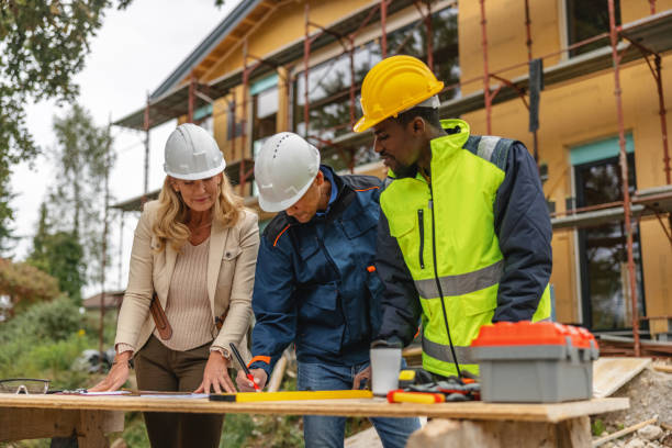 Senior female civil engineer and two male construction engineers wearing hard hats and safety vests standing at construction site and examining blueprints.