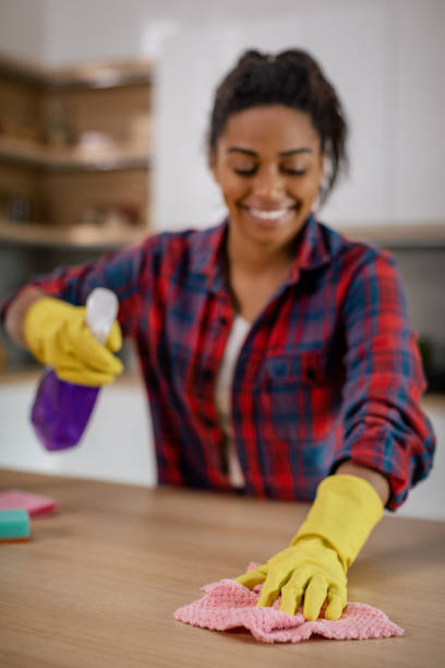 Cheerful young black lady wipes table from dirt with cleaning supplies in white kitchen interior, close up. Lifestyle, cleaning, household chores, hygiene and health care alone at home in spare time
